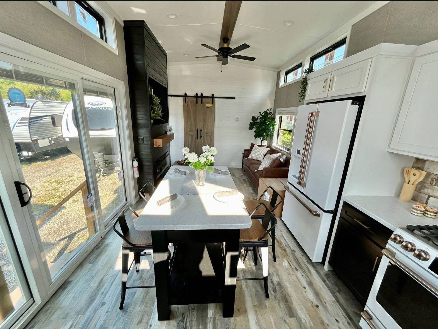 Modern tiny house interior with a bright kitchen, featuring an island with stools, white cabinets, a brown sofa, and large windows. Cozy and inviting.