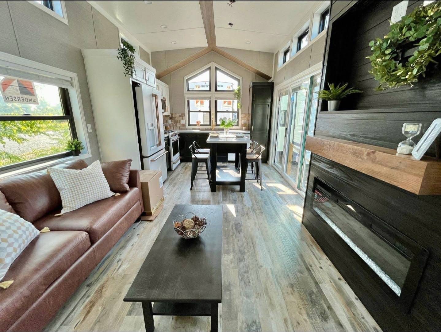 Modern tiny house interior with open layout. Features a cozy brown sofa, wood floors, kitchen with island seating, large windows, and a black fireplace with greenery. Bright and inviting atmosphere.