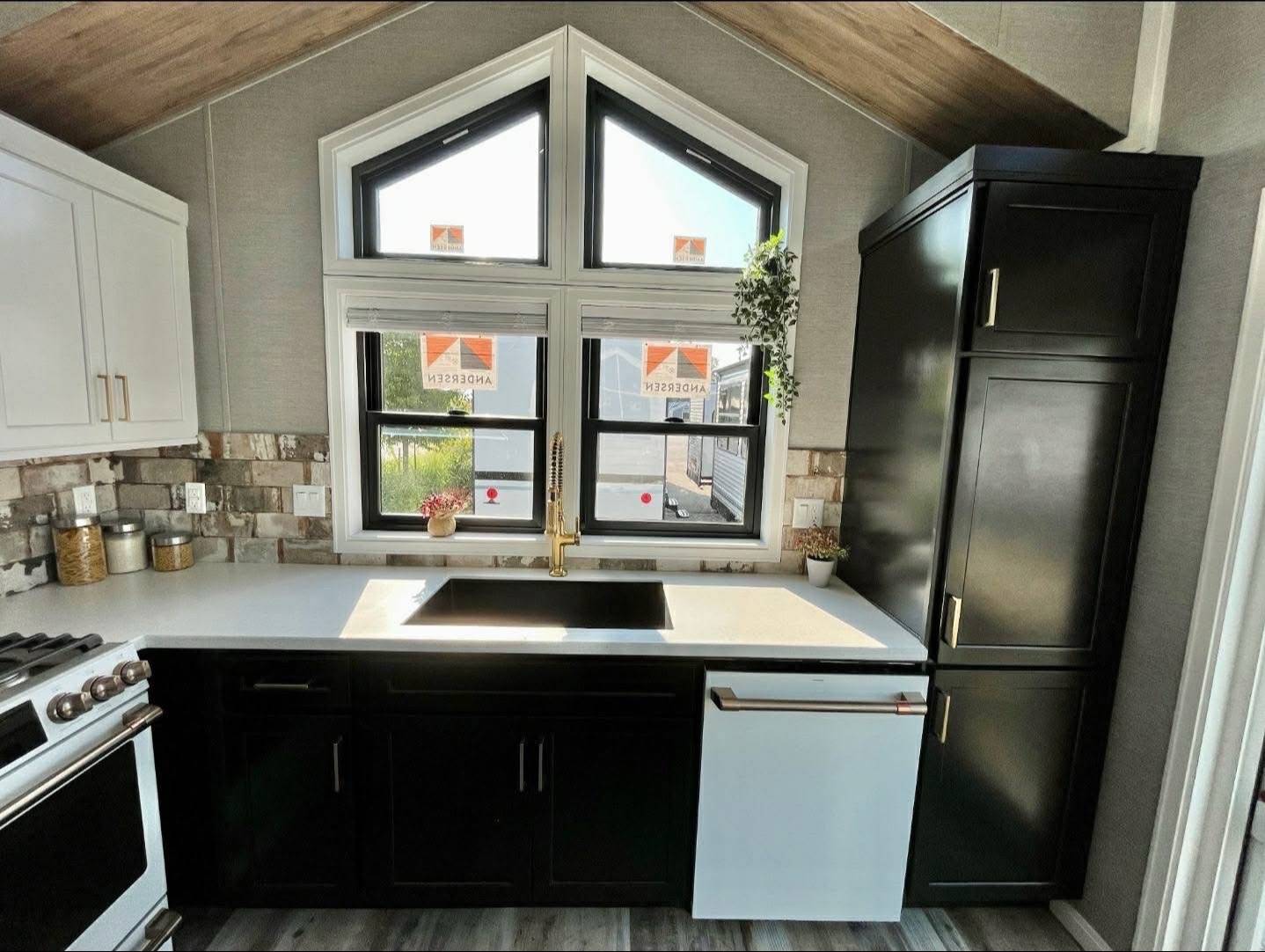Modern kitchen with black cabinets, white countertops, and a sleek golden faucet. Large windows let in natural light, creating a fresh ambiance.