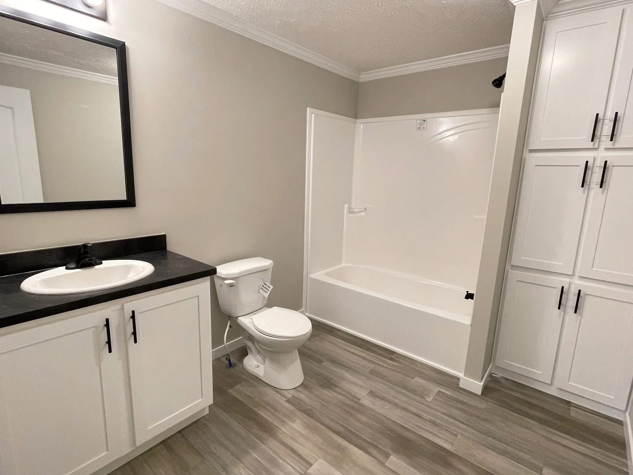 Modern bathroom with wood-style flooring, white cabinets, and black accents. Includes a bathtub, toilet, sink with a mirror, and ample storage.