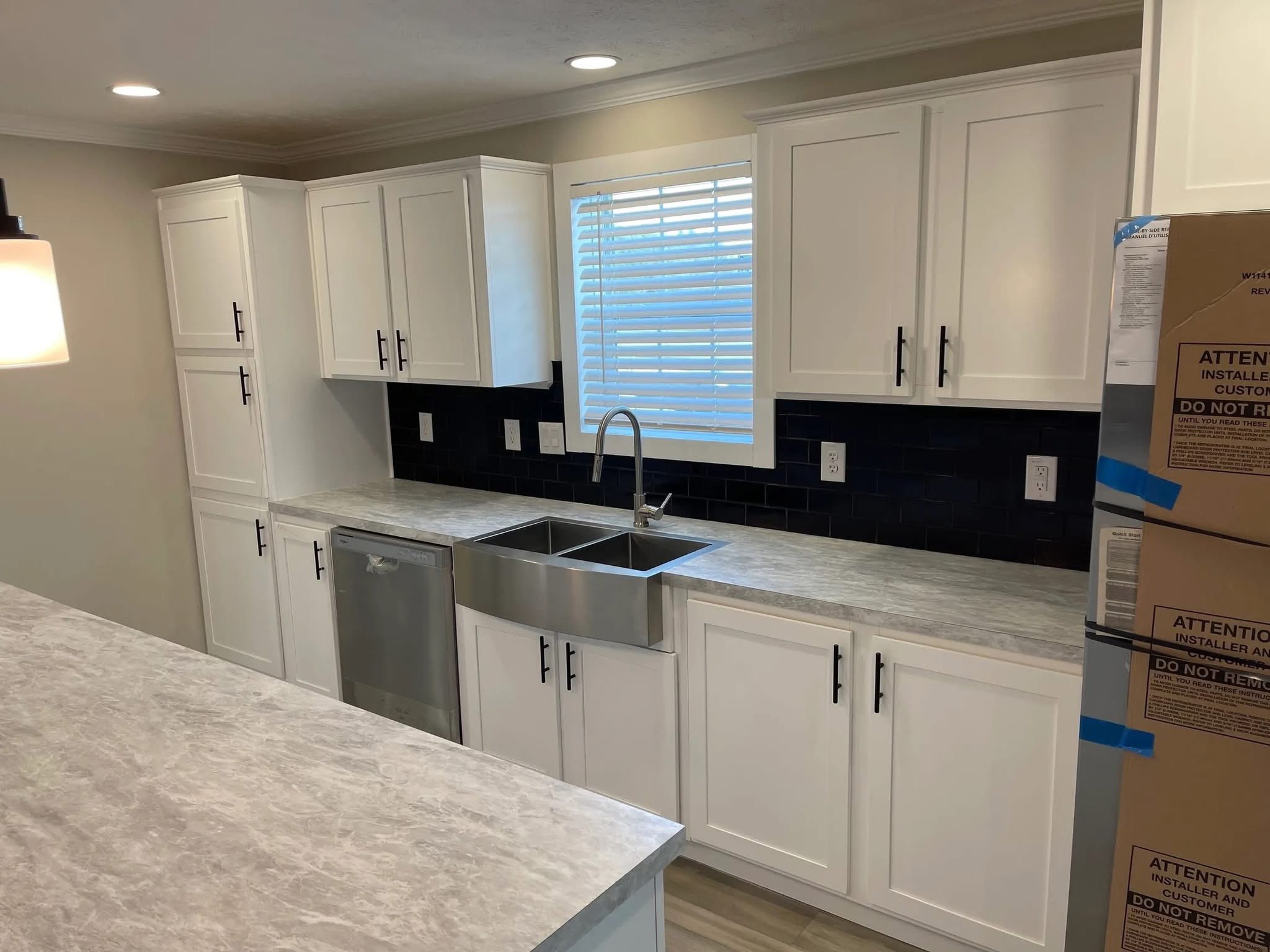 Modern kitchen with white cabinets, dark backsplash, and a stainless steel sink under a window with blinds. Boxes are stacked on the right.