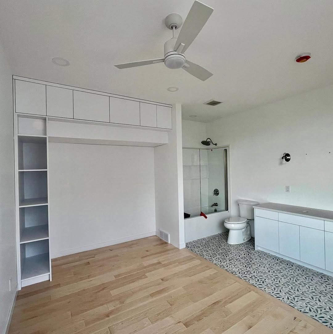 Minimalist room with light wood flooring and white walls. Features built-in shelves, modern ceiling fan, bathroom with patterned tiles, and white cabinetry.