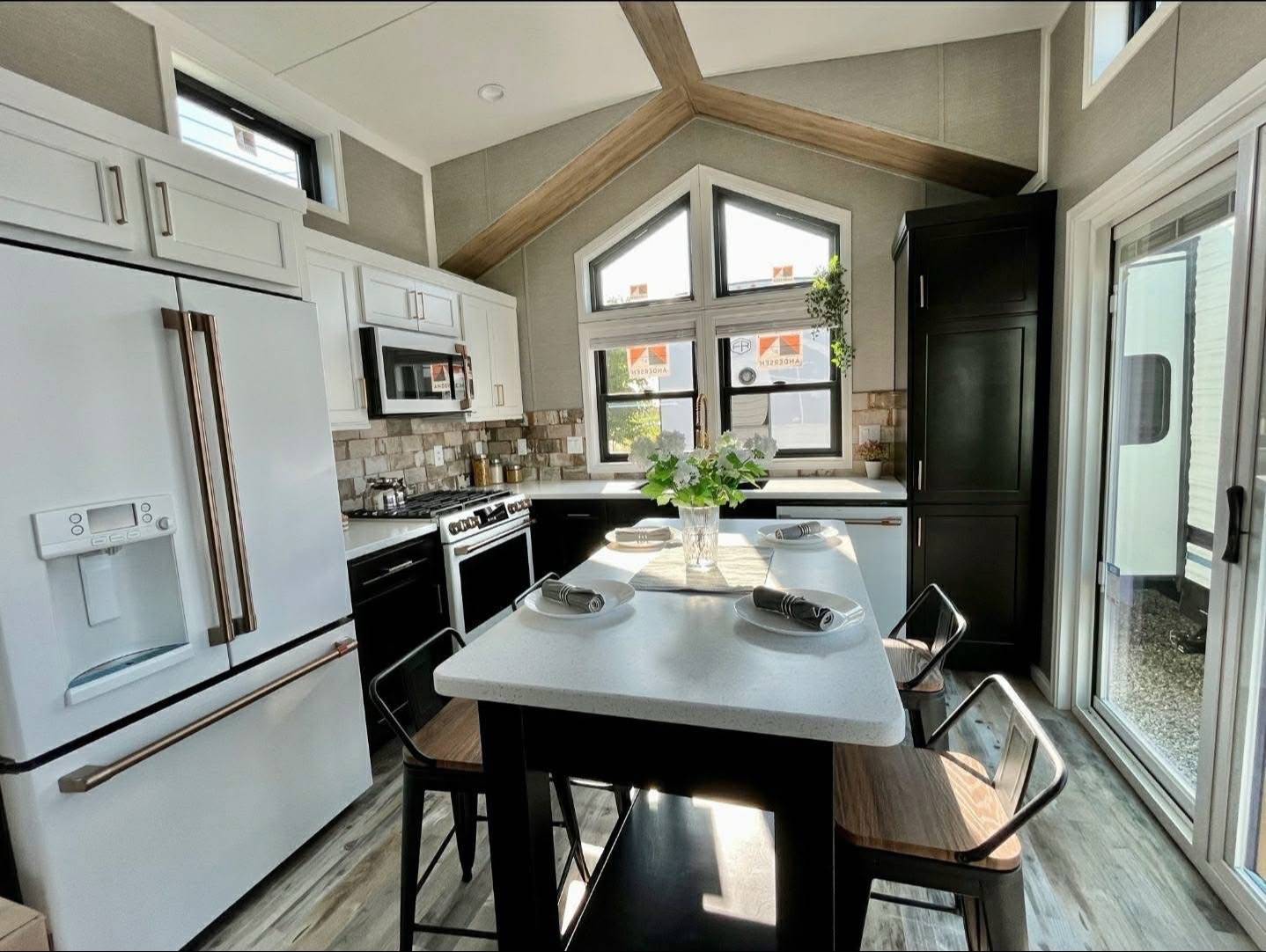 Modern kitchen with a high ceiling and large triangular window. Features white cabinets, an island with chairs, stainless steel appliances, and decorative plants, creating an inviting atmosphere.