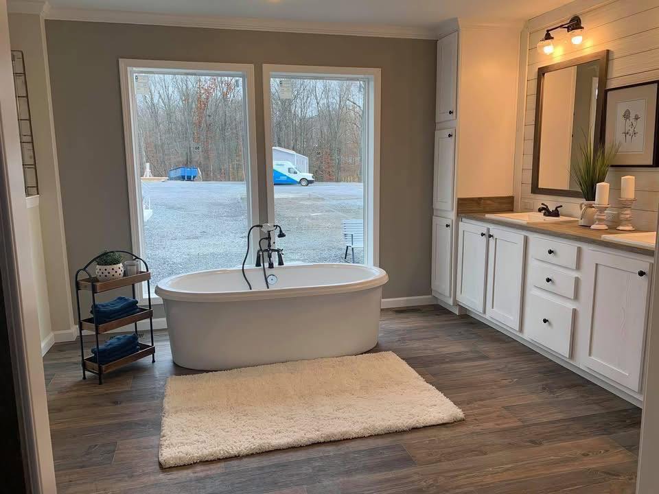 Modern bathroom with a freestanding tub on a beige rug, wood flooring, large windows showing outdoor scenery, white cabinets, and elegant decor.
