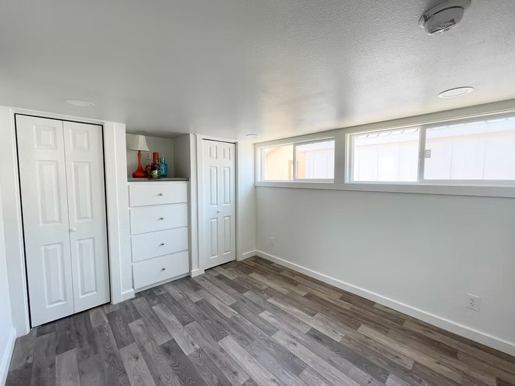 Minimalist room with gray laminate flooring, white walls, and a white dresser. Two closets flank the dresser. Horizontal windows let in natural light.