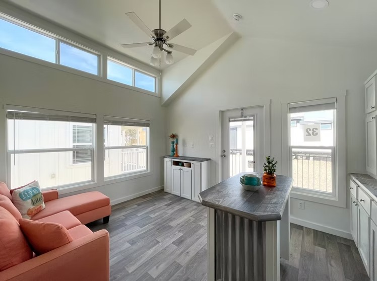 Modern living room with high ceilings and large windows. Features a coral pink sofa, gray wood floors, ceiling fan, and a central counter with colorful vases. Cozy and bright atmosphere.