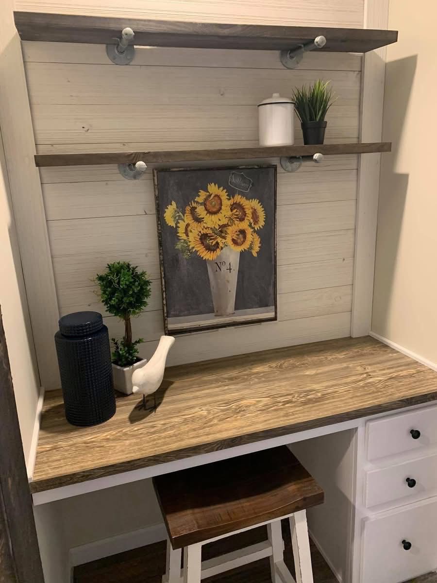 A cozy desk setup with a wooden tabletop, two wall shelves, a framed sunflower painting, and decor items like a potted plant and ceramic bird.