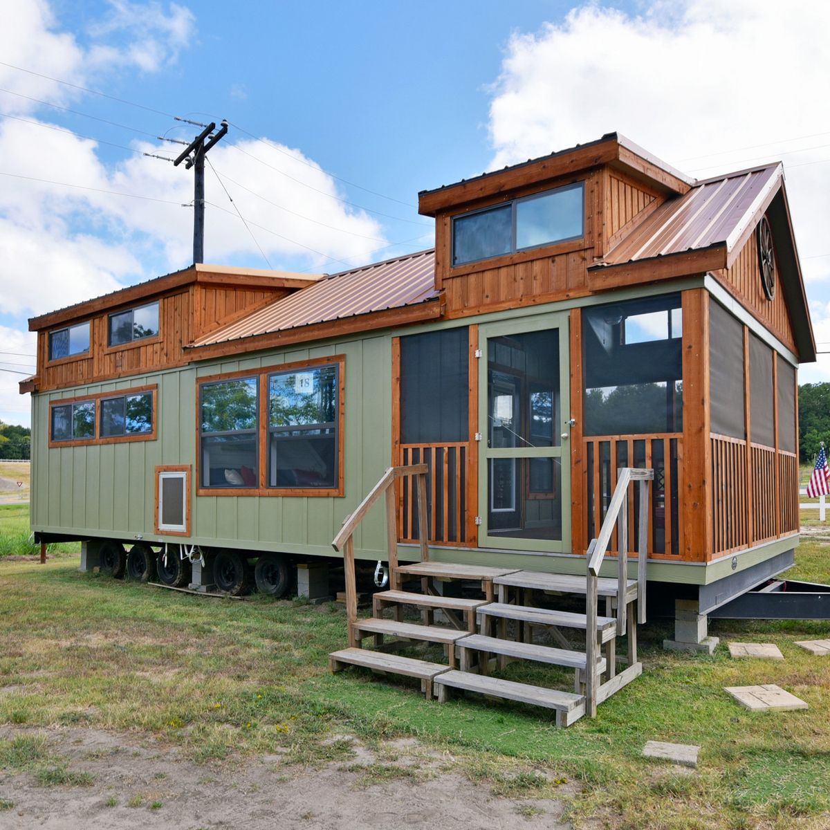 A tiny house on wheels with a rustic wooden exterior and green painted walls, set on grassy land under a blue sky with fluffy clouds.