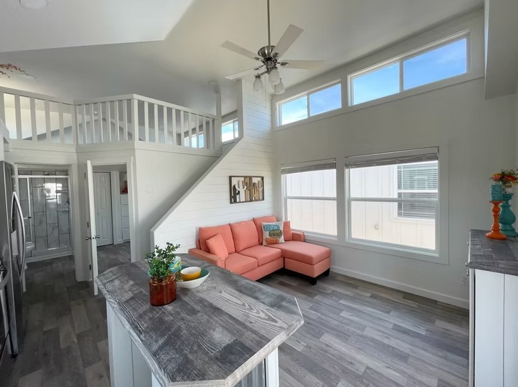 Bright, modern living room with high ceiling and large windows. Features a light coral sectional sofa, grey wood floors, and a loft area. Cozy and airy ambiance.