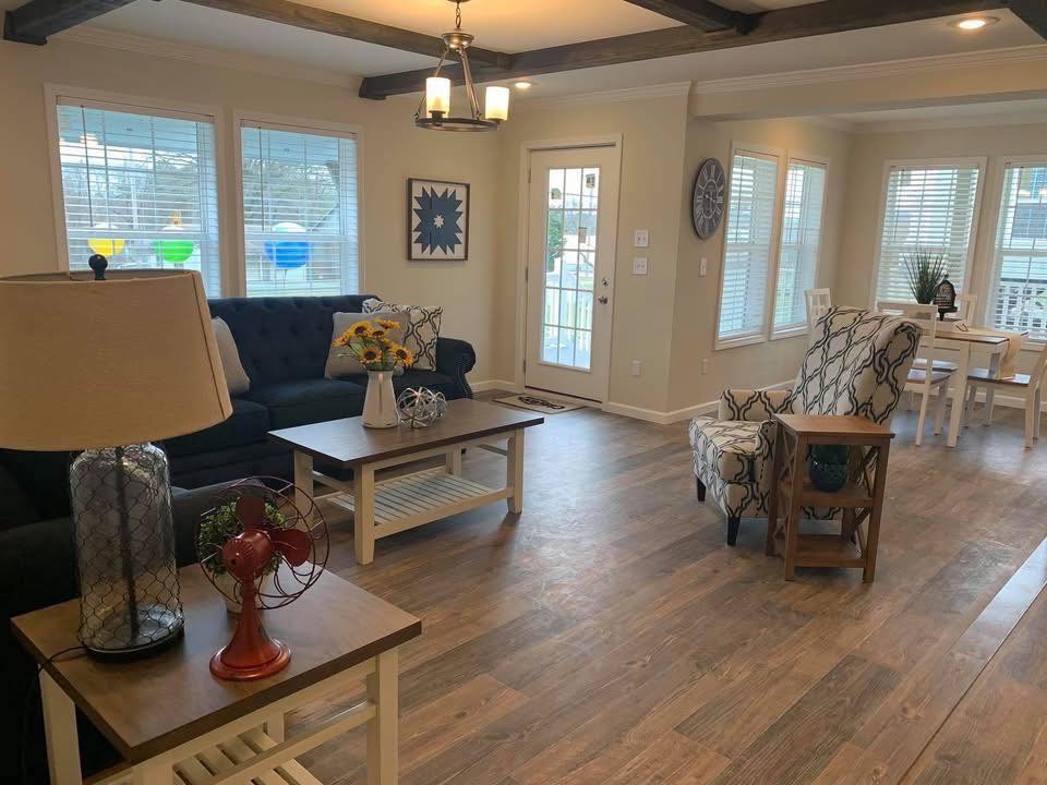 Warm living room with hardwood floors, a dark couch with sunflower centerpiece on coffee table, patterned armchair, and dining area by large windows.