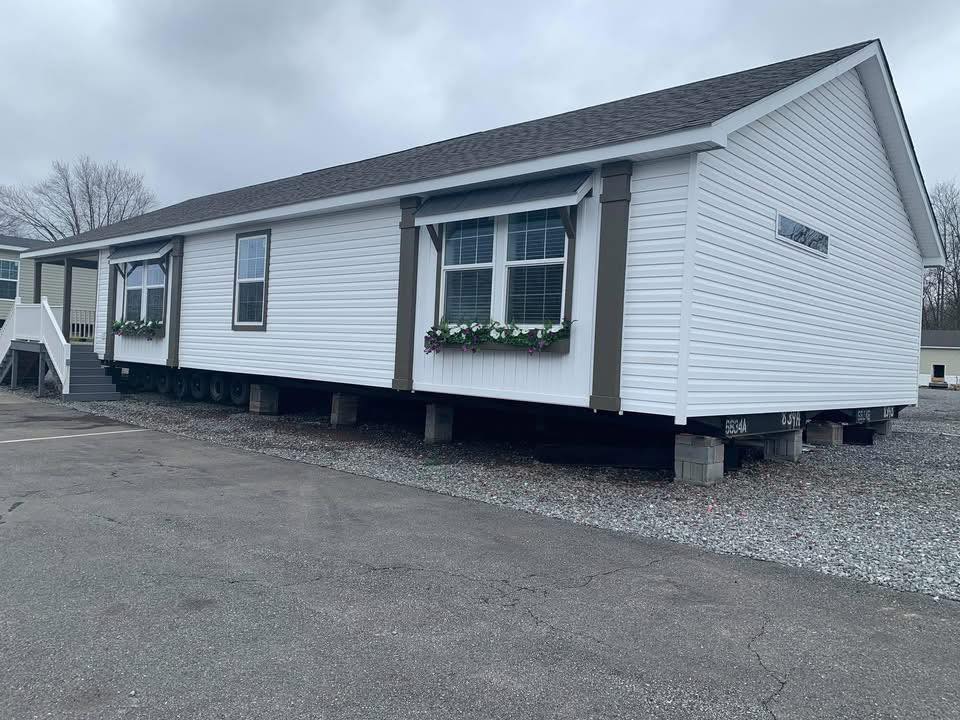 A large mobile home on blocks with white siding and dark trim, featuring flower boxes beneath wide windows, sits on a gravel lot under a cloudy sky.