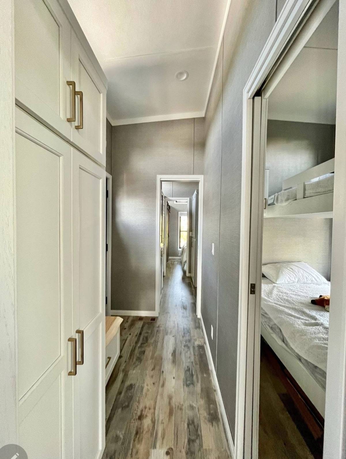 Narrow hallway with wood flooring and white cabinets on the left. On the right, a bedroom with bunk beds visible. Bright natural light fills the space.