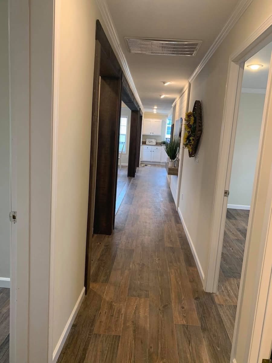 Narrow hallway with wooden floor leading to a well-lit kitchen. Walls are beige, adorned with a sunflower wreath, and a green plant adds freshness.