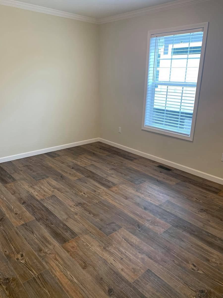 An empty room with warm beige walls and wooden flooring. A large window with white blinds lets in soft natural light, creating a calm ambiance.