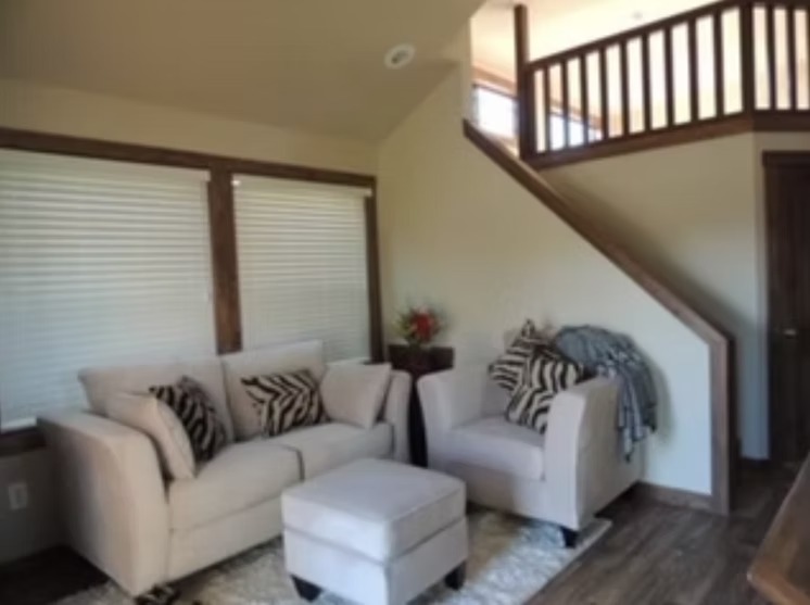Cozy living room with beige sofas and zebra-patterned cushions. A footstool sits in front of the sofas on a plush rug. Wooden stairs lead to an upper level.