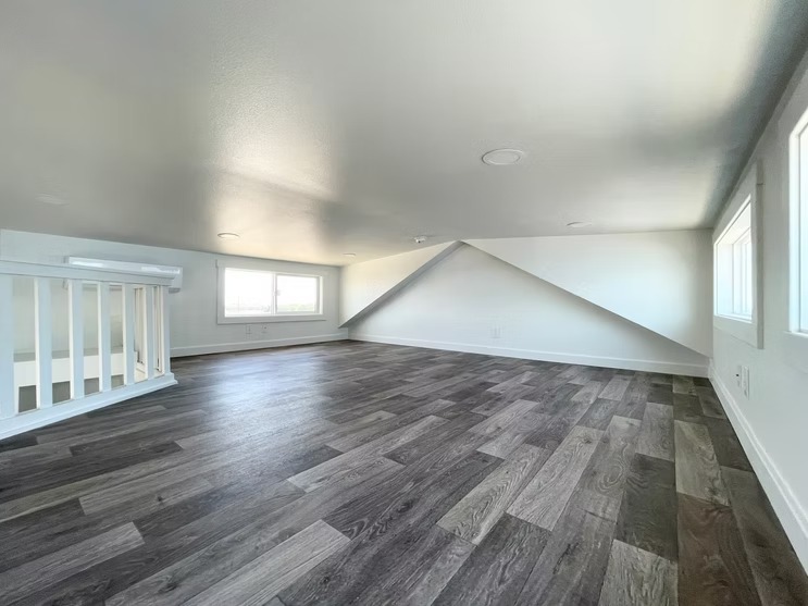 Spacious attic with dark wood flooring, white walls, and sloped ceiling. Natural light streams in from small windows, creating a cozy ambiance.