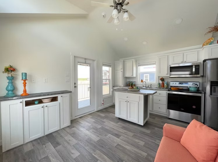 Bright kitchen with white cabinets, wooden flooring, and stainless steel appliances. Features vibrant decor, an island, and a cozy orange couch.