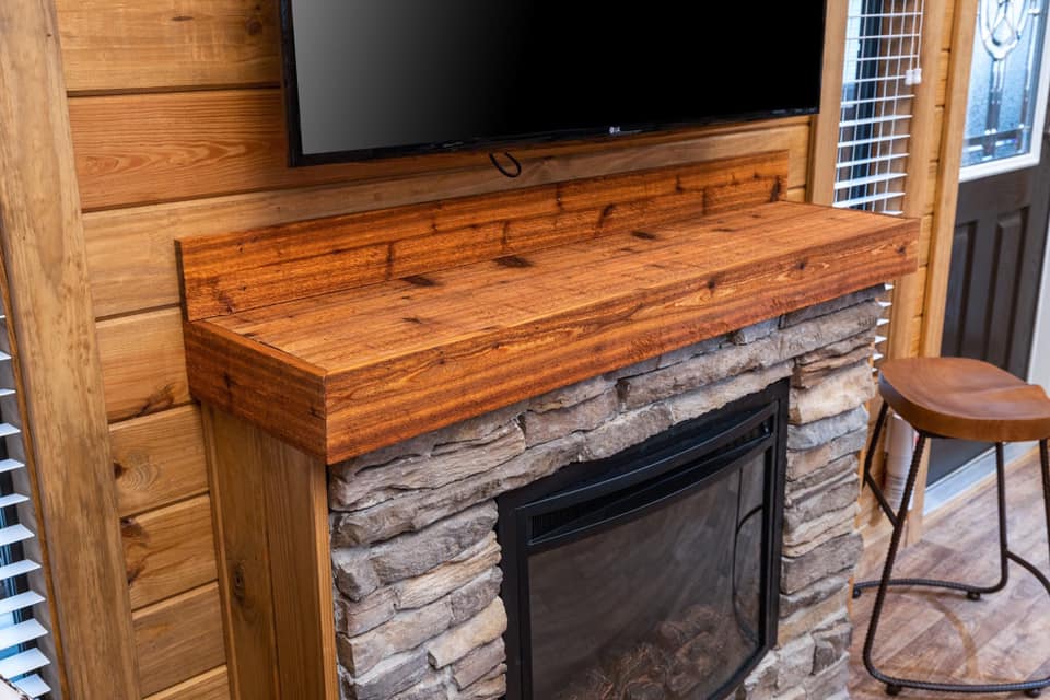 Wood-paneled room with a stone fireplace and wooden mantel. A flat-screen TV is mounted above. A brown stool rests on the wooden floor nearby. Warm and cozy ambiance.