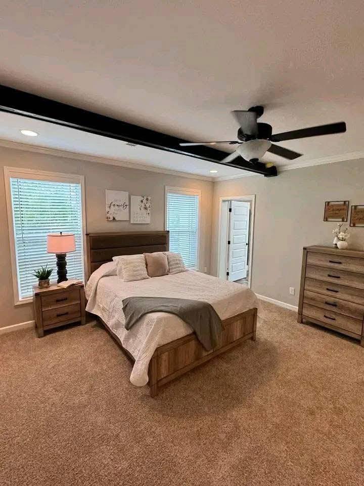 Cozy bedroom with a wooden bed, beige bedding, and a gray blanket. Two windows, a ceiling fan, nightstand with lamp, and dresser enhance warmth.