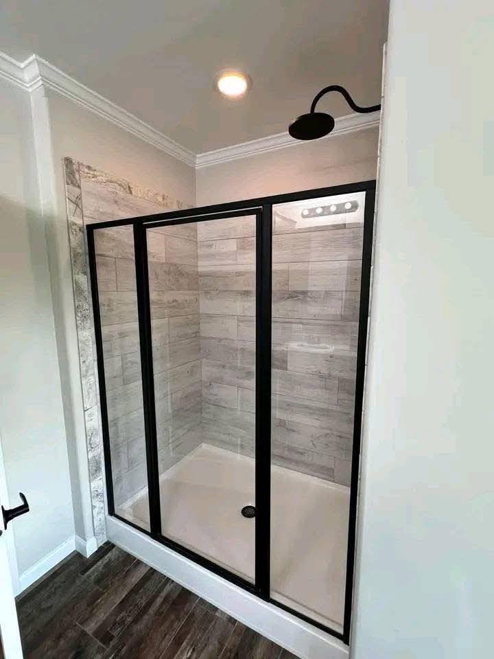 Modern shower with black-framed glass doors, light wood-style tiles inside, and dark wood flooring outside, under soft ceiling lighting.