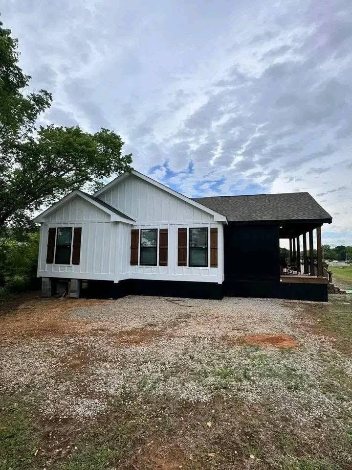 A modern small house with a white facade and brown shutters stands beside a black porch. It's set against a cloudy sky, surrounded by trees and gravel.