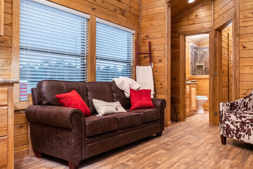 Cozy living room with wooden walls and floor. A brown sofa with red and white pillows is near two large windows. A bathroom is visible in the background.