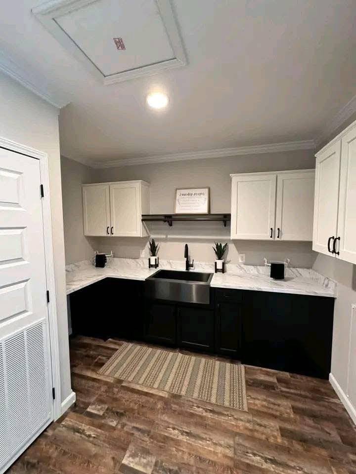 Modern kitchen with black lower cabinets, white upper cabinets, and marble countertops. There's a farmhouse sink, two small potted plants, and a light wood floor. The tone is clean and minimalist.