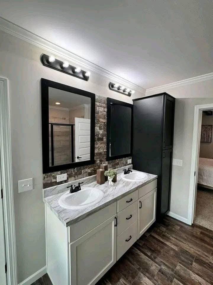 Modern bathroom with dual sinks on a white vanity, black-framed mirrors, stone backsplash, and soft lighting. Features wood-style flooring, black cabinet.