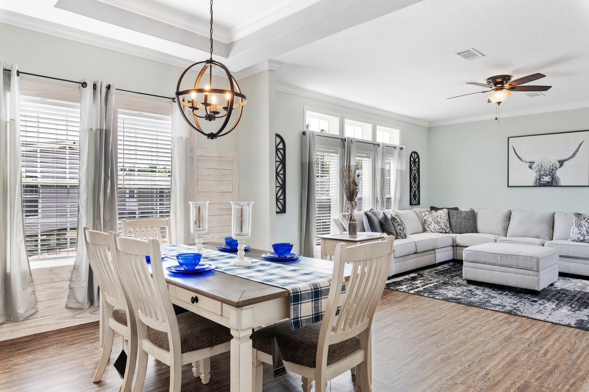 A modern, cozy living and dining space featuring a wooden table with blue dishware, a gray sectional sofa, and a bull artwork. Light filters through large windows.