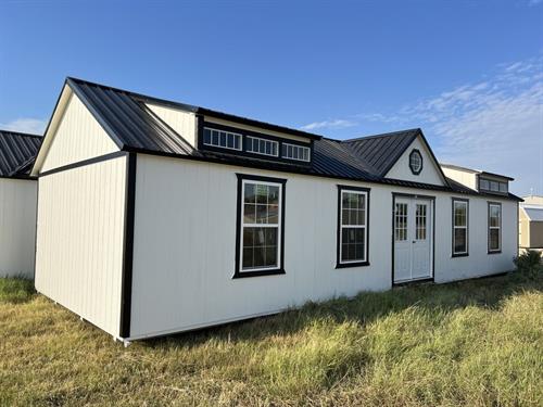 A white modular cabin with black trim sits on grassy ground under a clear blue sky. It has a metal roof, multiple windows, and a double door.