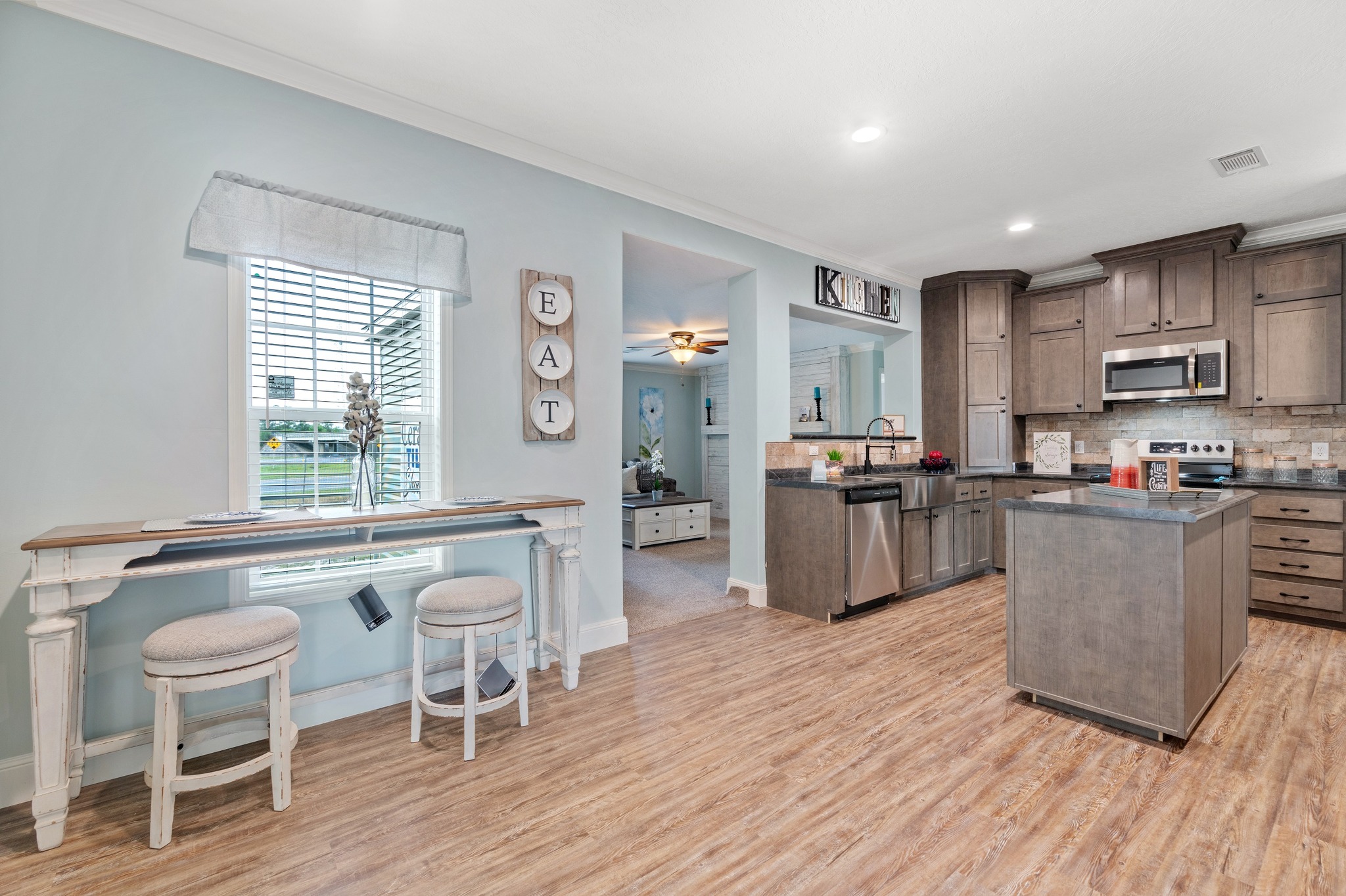 Bright kitchen with wooden floors and light blue walls. Dark wood cabinets, stainless steel appliances, and an island create a cozy ambiance. A wall sign reads "EAT."