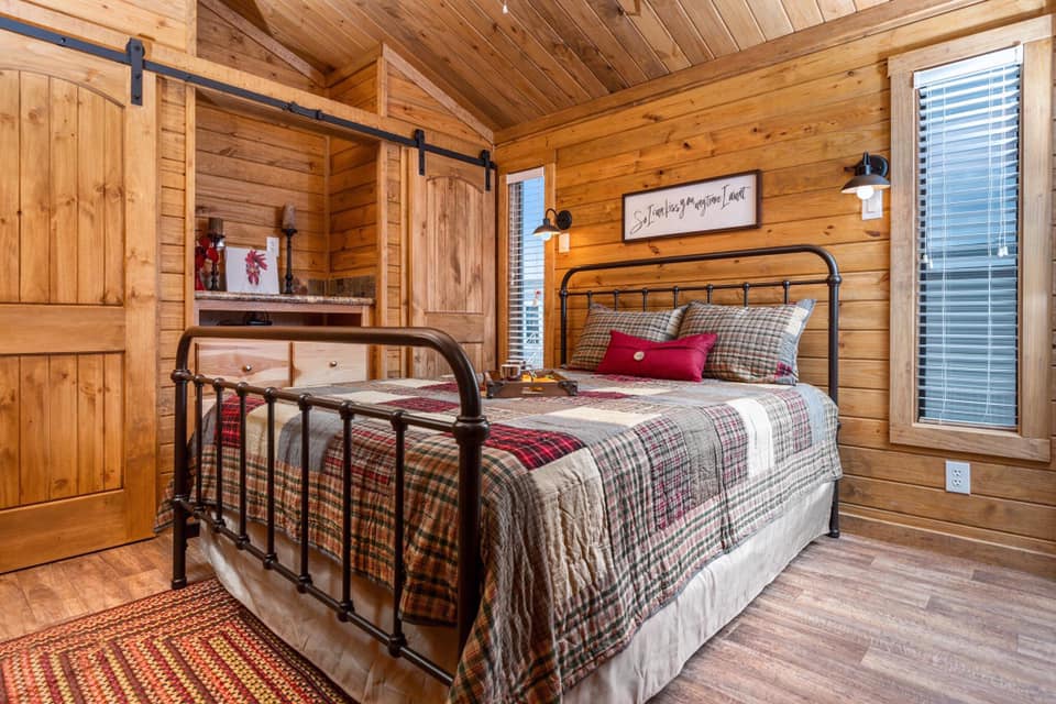 Cozy rustic bedroom with wooden walls and ceiling, featuring a metal-framed bed adorned with plaid bedding. Warm lighting and barn-style closet doors.