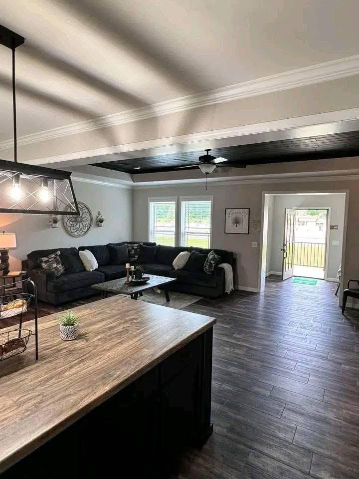 Spacious living room with wooden flooring, a dark L-shaped couch, and black ceiling accents. Sunlight streams through two windows near the couch.