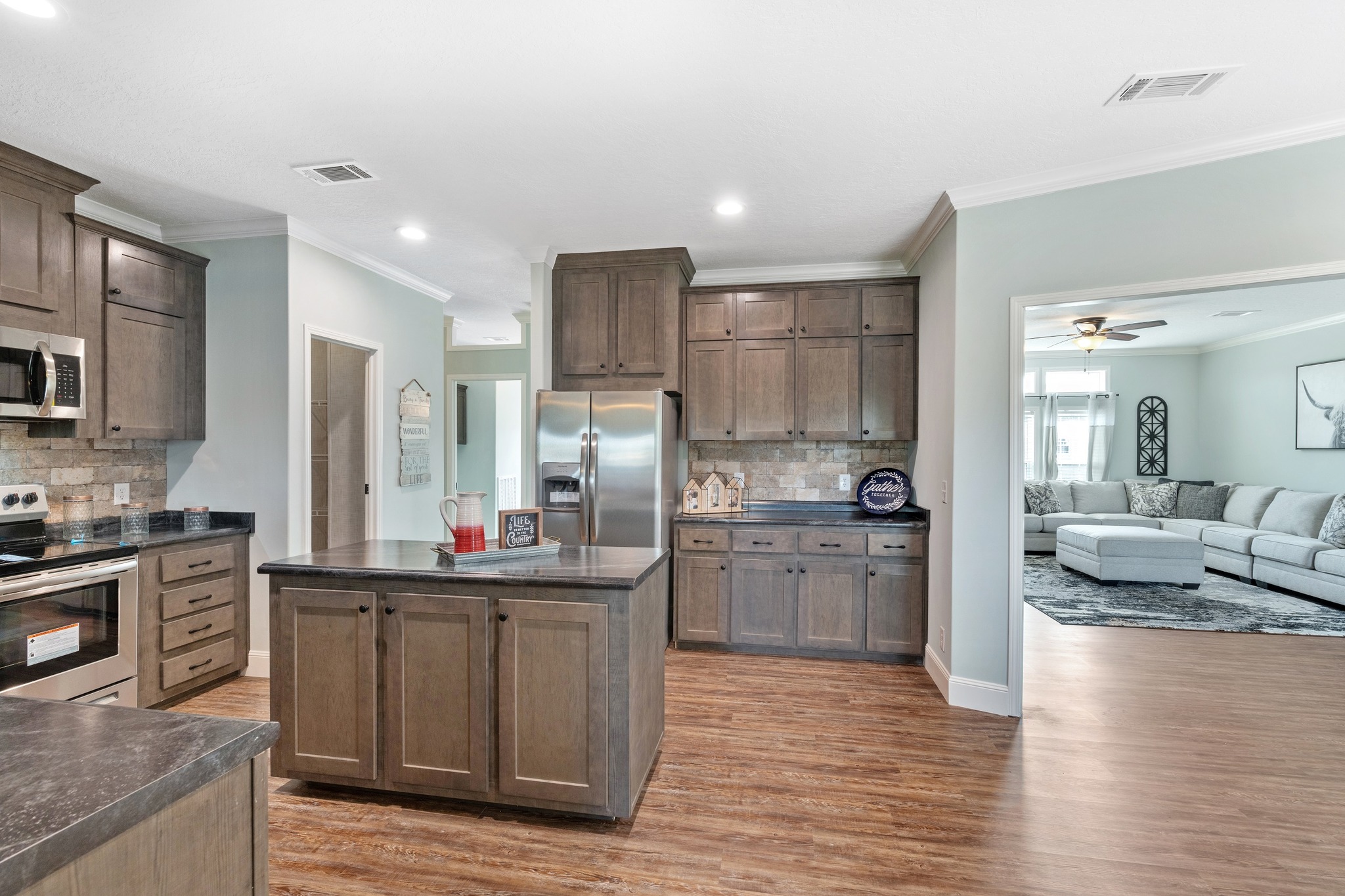 Spacious kitchen with wooden cabinets, dark countertops, and a central island. Stainless steel appliances and a glimpse of a cozy living room.