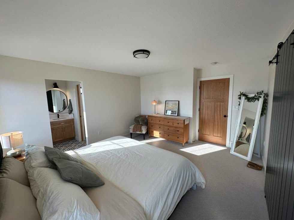 A cozy bedroom with a neatly made bed, wooden dresser, and soft lighting. A mirror leans against the wall, adding warmth to the bright, neutral space.