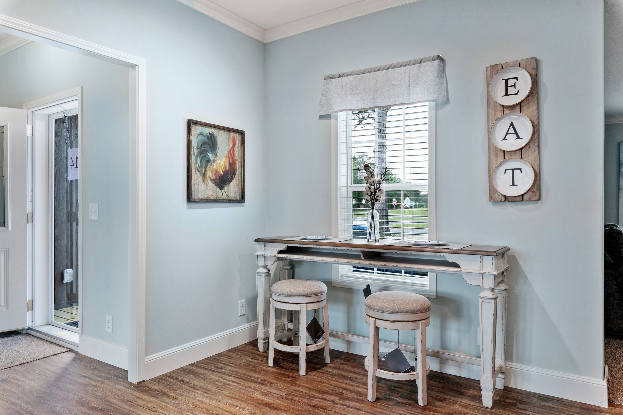 A cozy interior corner features a light blue wall, a vintage table with two stools, a rooster painting, and "EAT" decor. The window overlooks greenery, creating a calm and welcoming atmosphere.