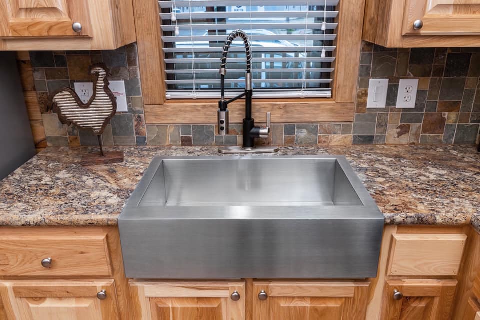 Modern kitchen sink with a stainless steel apron front on a granite countertop. Wooden cabinets, tile backsplash, rooster decor, and blinds.