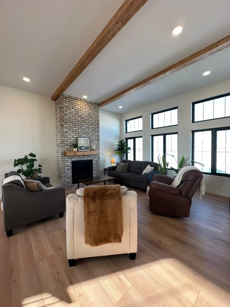 Spacious living room with high ceiling, wooden beams, and large windows. Cozy seating surrounds a brick fireplace. Bright, airy, and inviting.