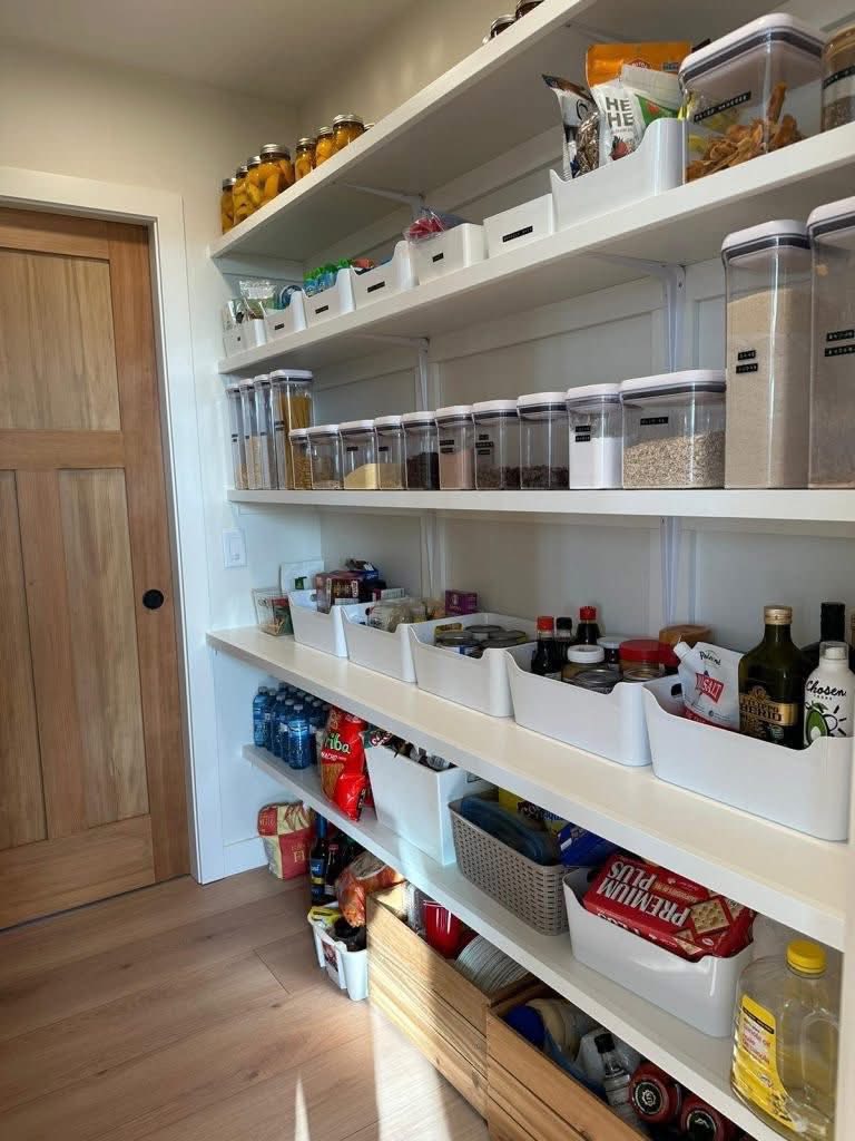 Organized pantry with labeled jars of dry goods on upper shelves, bins of snacks and sauces on middle shelves, and bottled drinks on the lower shelf. Warm, tidy ambiance.