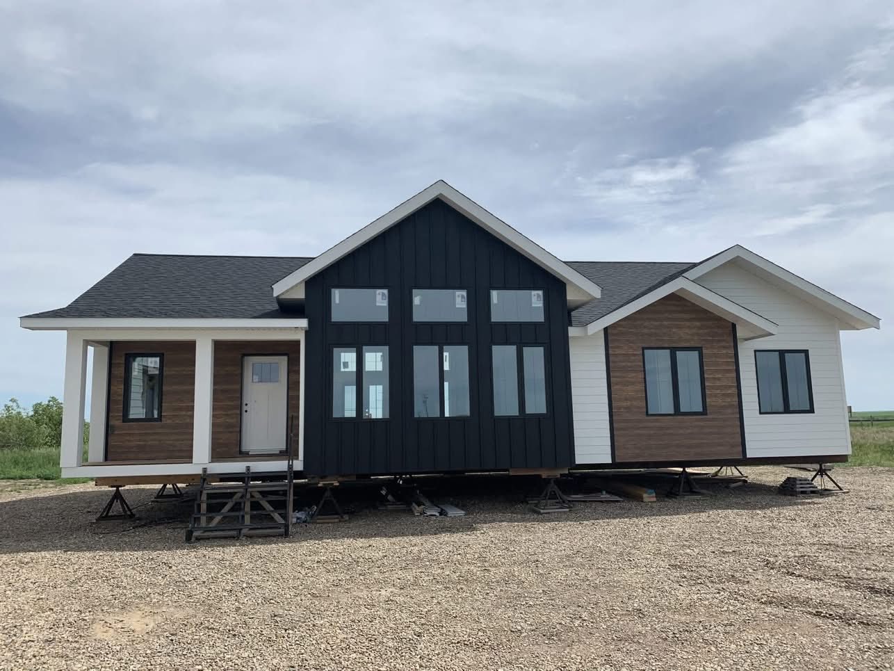 Modern modular home on stilts, featuring a gabled roof. Exterior combines black, white, and wooden panels, set against a cloudy sky backdrop.