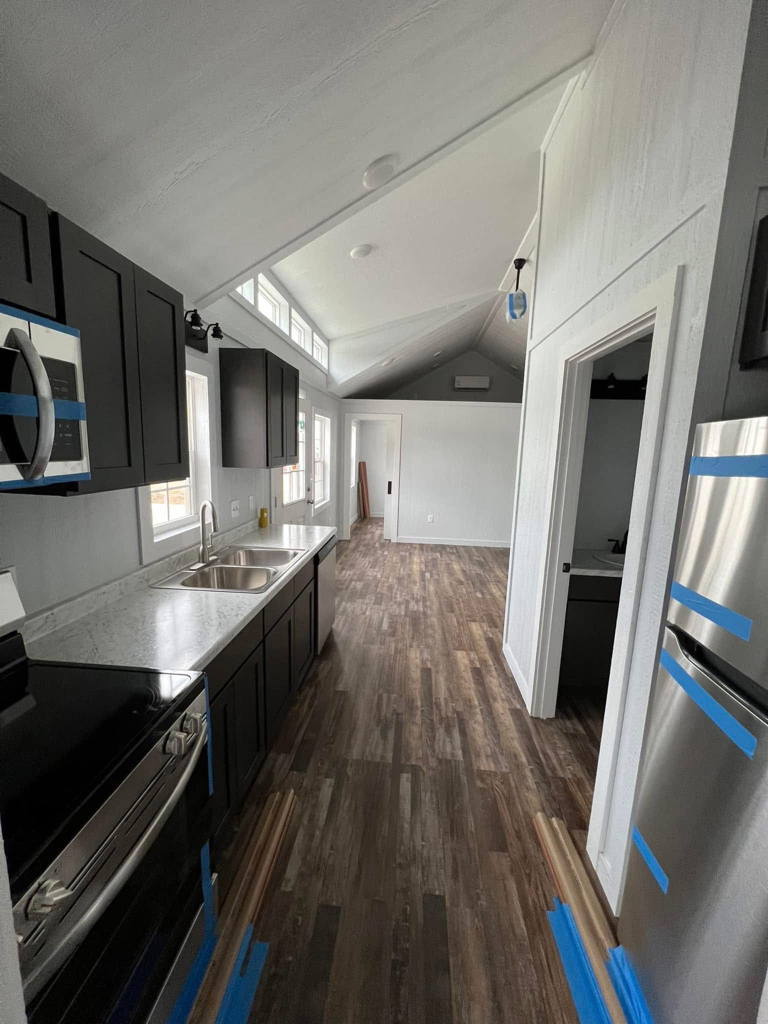 Modern, narrow kitchen with dark cabinets, white marble countertops, a stainless steel sink, and wood flooring. Bright windows add natural light.