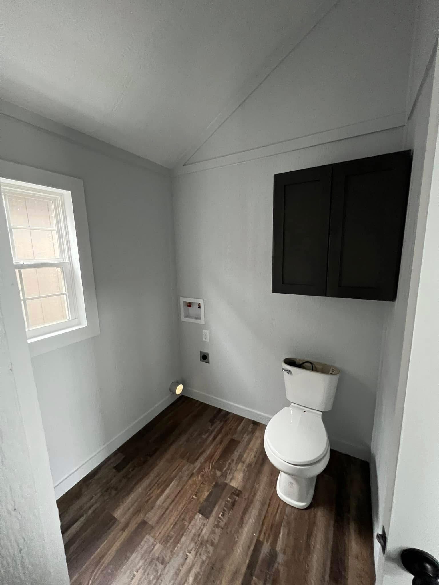 Small bathroom with wooden floor, featuring a white toilet, black cabinet, and a single window. The space is minimal and well-lit.