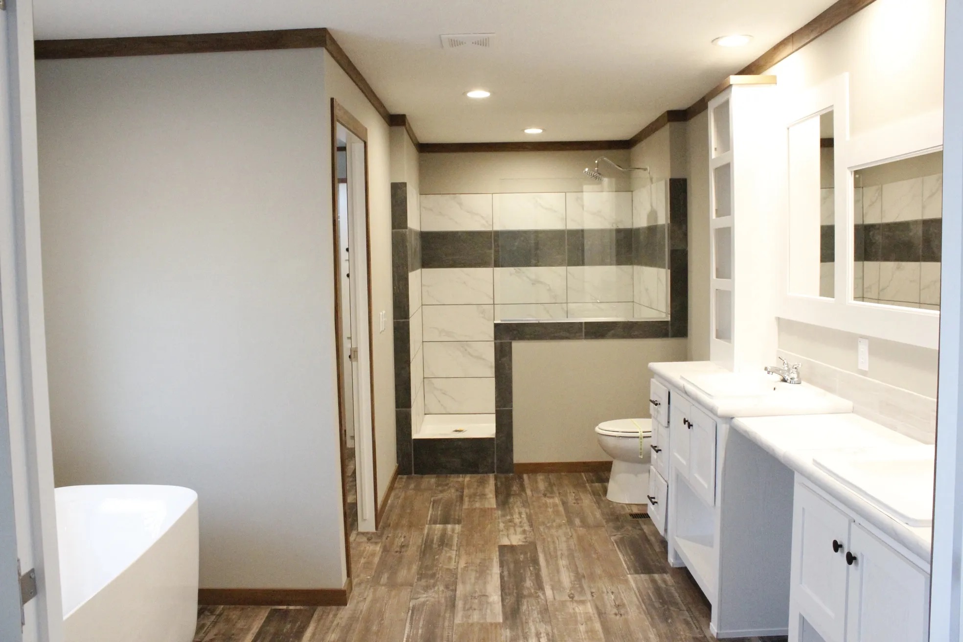Modern bathroom with wood flooring, featuring a spacious shower with gray and white tiles, a white vanity with dual sinks, and soft lighting.