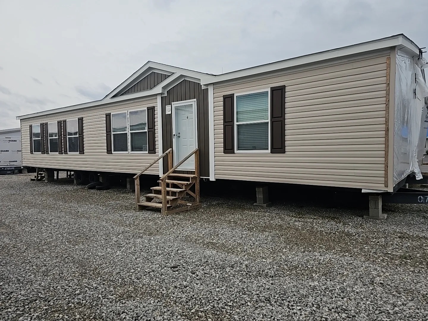 A beige double-wide mobile home on a gravel lot, featuring brown shutters, a white door with steps, and a gabled roof, conveying a simple, residential tone.