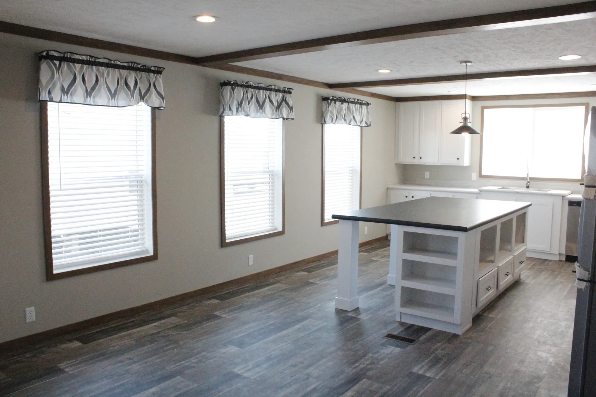 Spacious, modern kitchen with wood floors, three large windows with patterned valances, white cabinets, and a central island under pendant lighting.