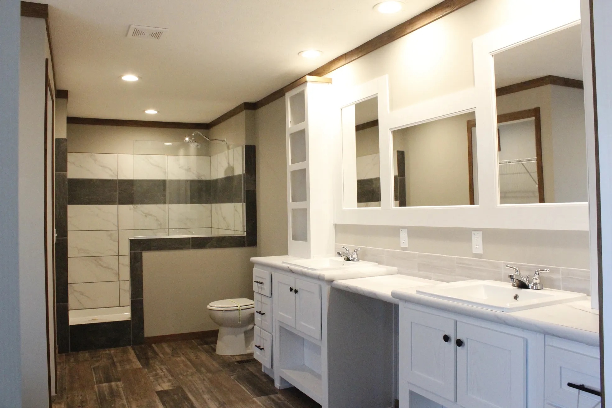 Modern bathroom with wood flooring, two sinks, and large mirrors on a dual vanity. A walk-in shower is visible in the background. Soft, neutral tones convey a clean, spacious feel.
