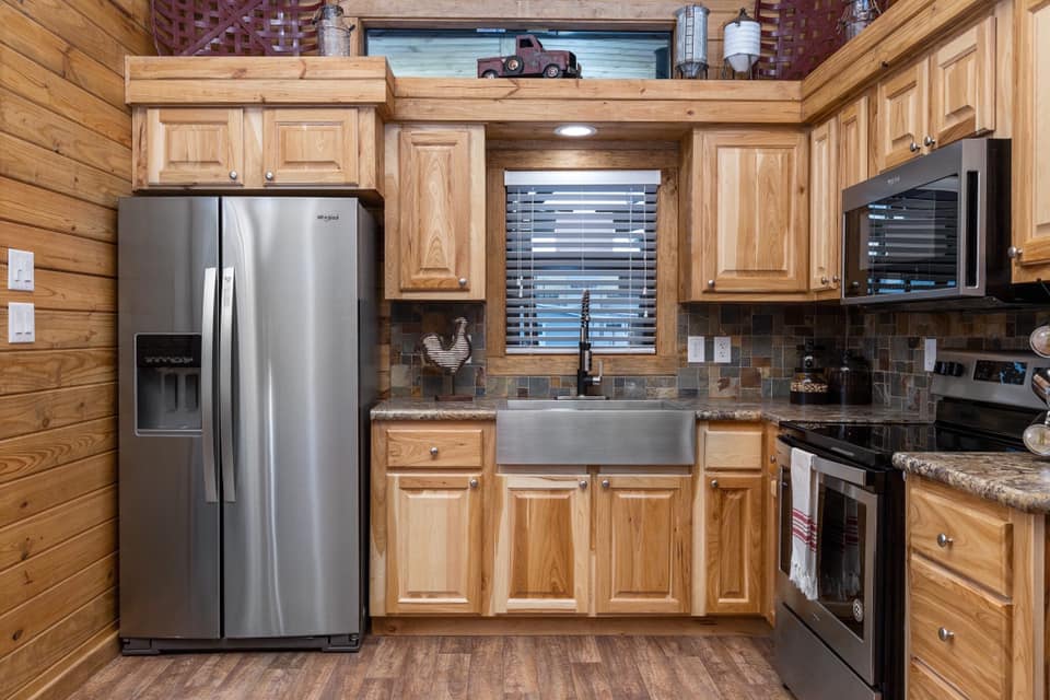 Rustic kitchen with wooden cabinets, stainless steel appliances, and a farmhouse sink. A small window with blinds adds natural light. Cozy and warm atmosphere.