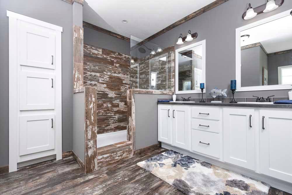 Modern bathroom with white cabinetry, rustic wood accents, gray walls, and dual mirrors. Features a walk-in shower and a patterned rug for a cozy feel.