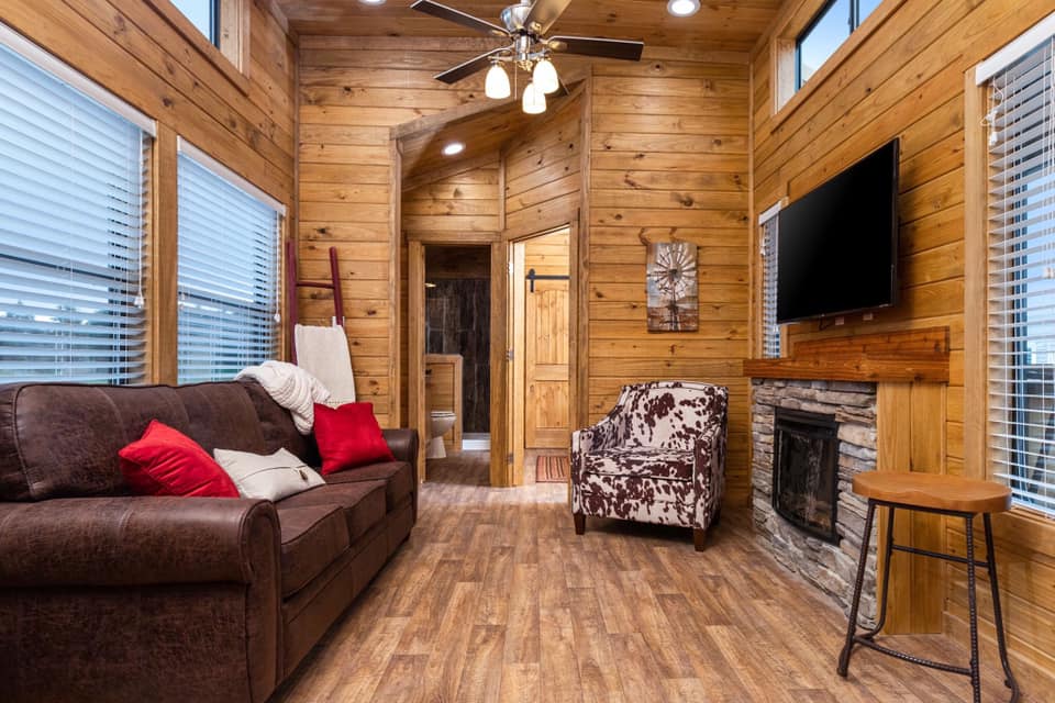 Cozy living room with a wooden interior, featuring a brown sofa with red cushions, a patterned armchair, a stone fireplace, and a wall-mounted TV.