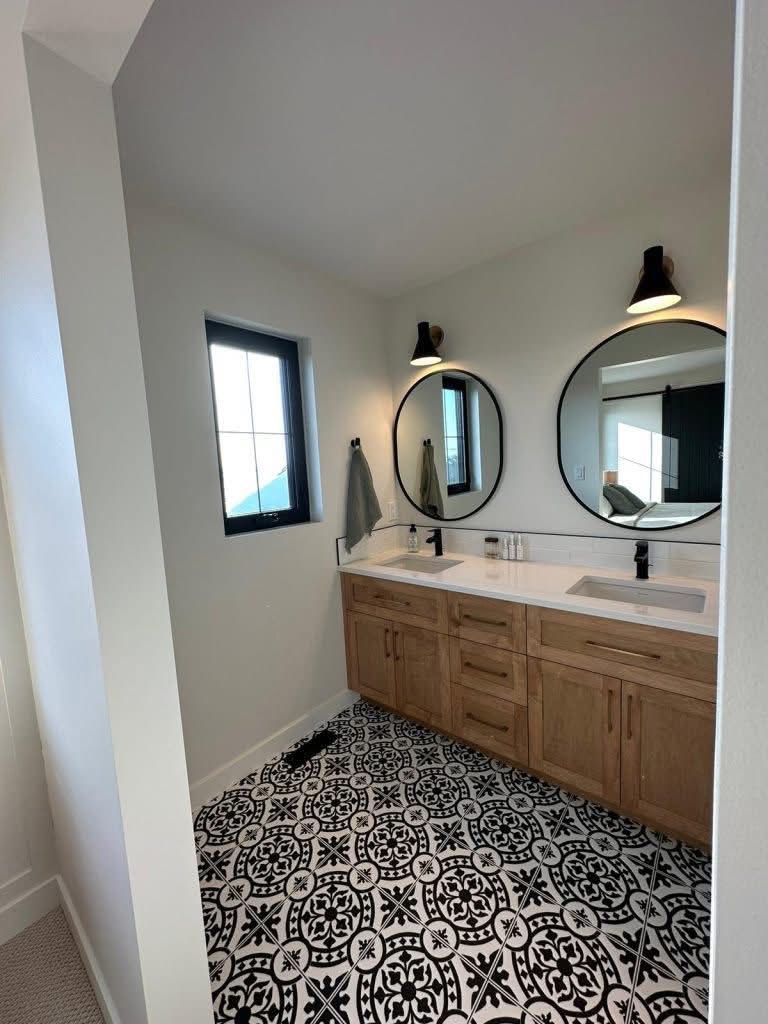 A modern bathroom features dual sinks with wooden cabinetry, round mirrors, and black sconces. The patterned black-and-white tile floor adds elegance.